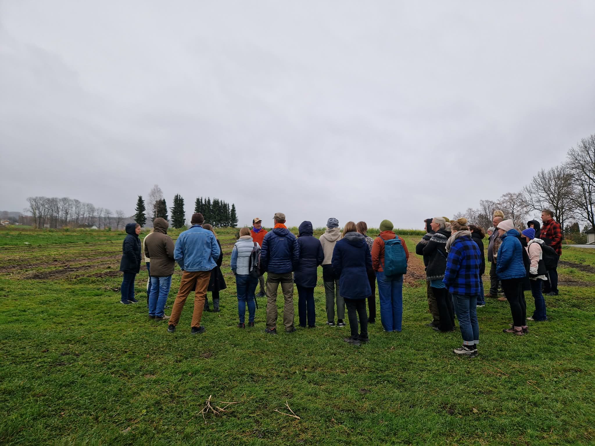 Engasjerte mennesker står i ring rundt mann som snakker om grønnsakshagen i bakgrunn. Det er på senhøsten så lite grønt å se.